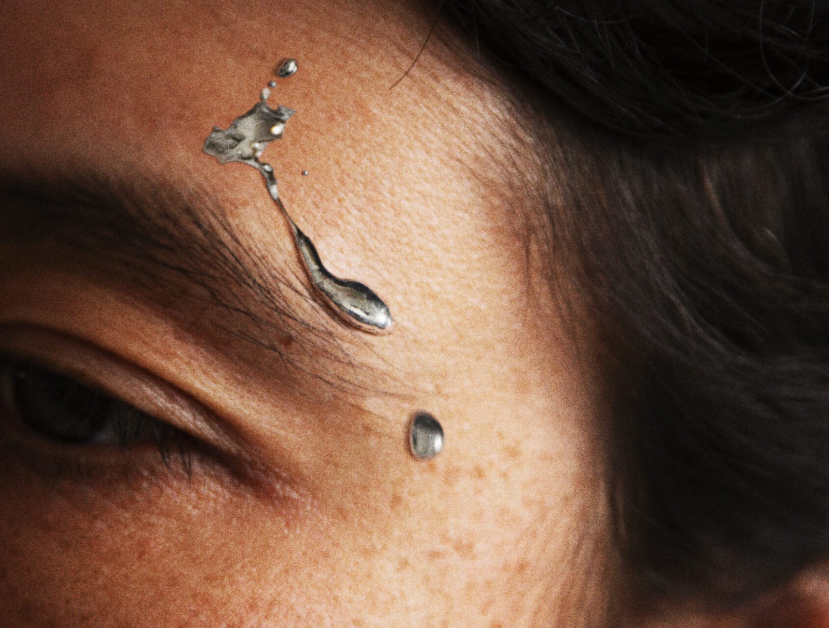 Image description: A very close up photo of a face with droplets of silver metal above the eyebrow.
